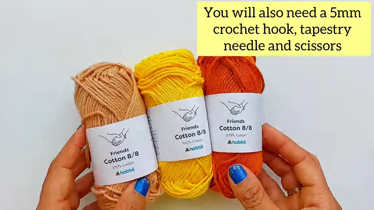 Hands holding beige, yellow, and orange cotton yarn beside a note listing a 5 mm hook, tapestry needle, and scissors