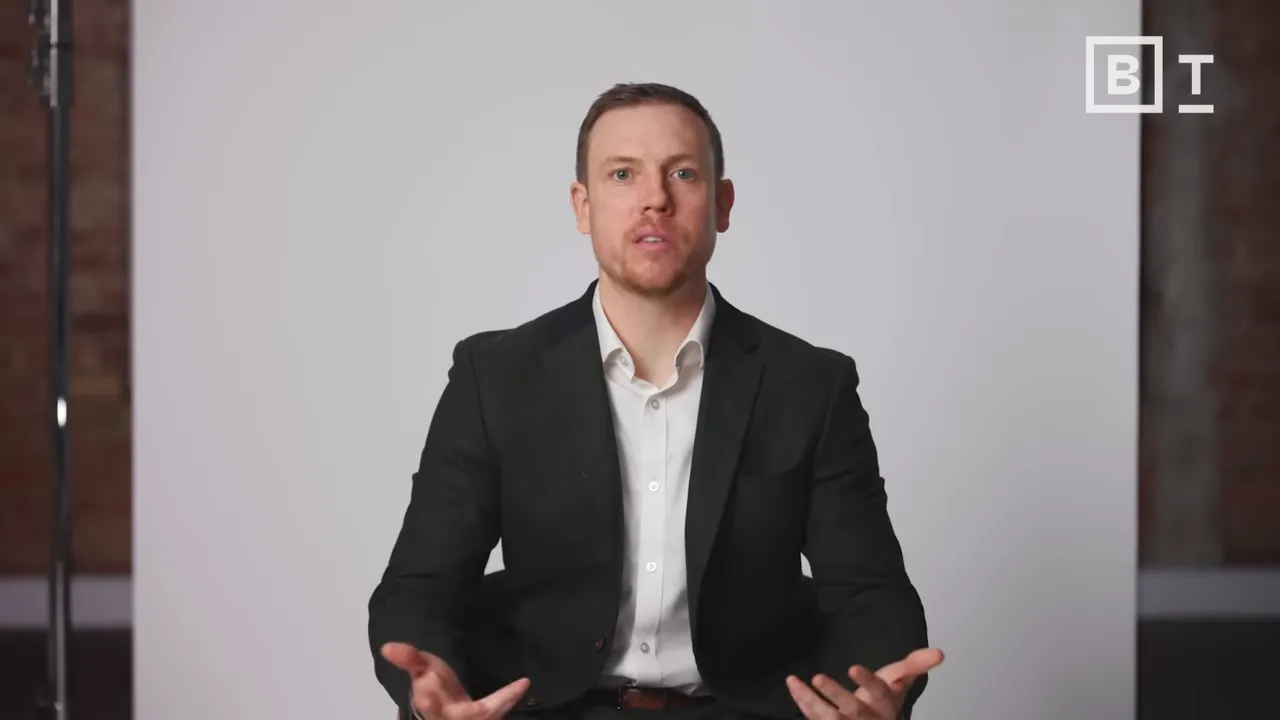 male speaker mid-shot, seated and gesturing with both hands against a neutral studio backdrop