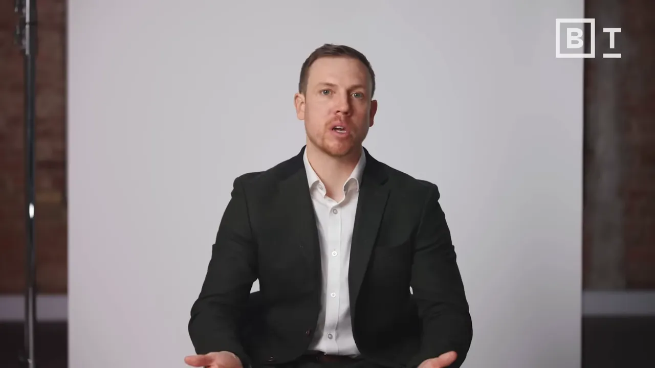 Presenter in suit, seated mid-gesture, clear studio lighting and backdrop.