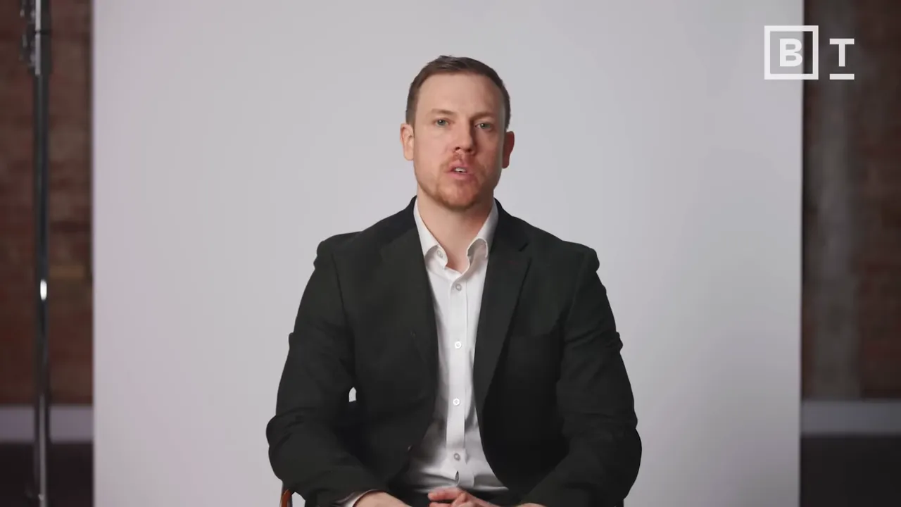 Man in suit speaking on camera, mid shot, neutral backdrop — clear, well-framed.