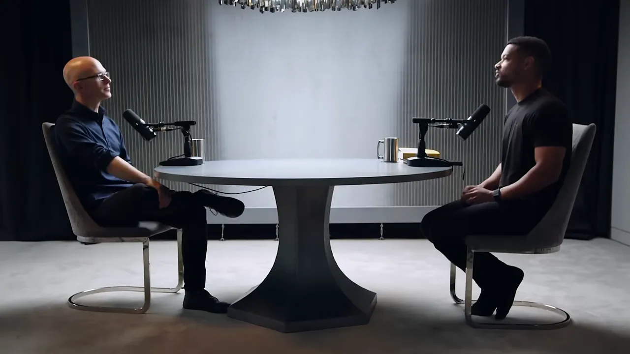 Two hosts seated across a round table with microphones in a minimalist studio setting.