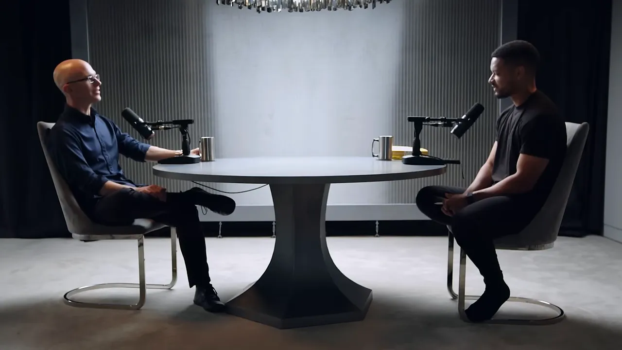 Two people sitting at a round table recording a podcast with microphones, studio backdrop