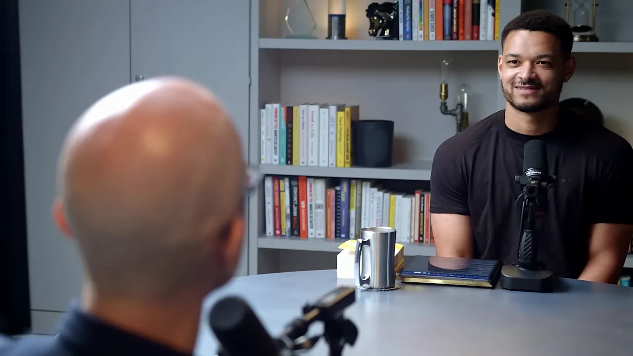 Two people in a podcast studio talking across a table with bookshelves and a microphone visible