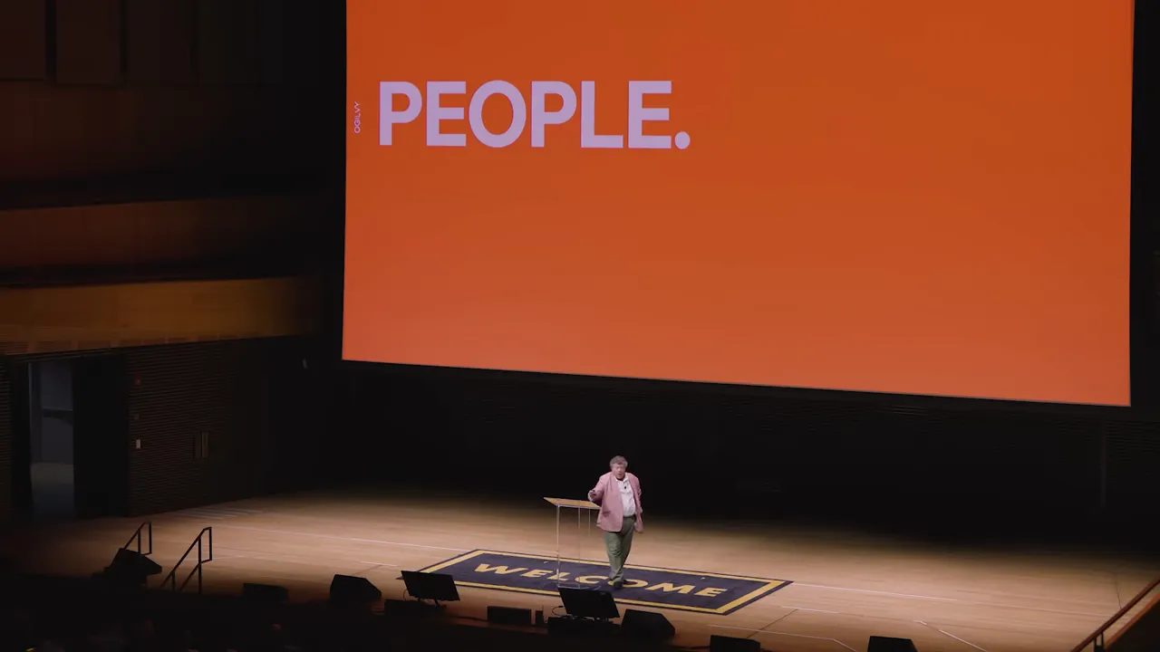 Auditorium stage with a large orange presentation slide reading 'PEOPLE.' and a presenter standing on stage