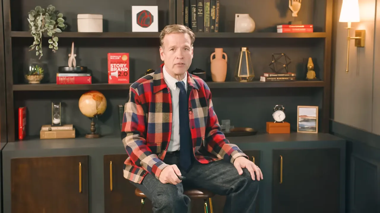 Presenter seated on a stool in front of a bookshelf-backed studio set, speaking to camera with hands resting on knees.