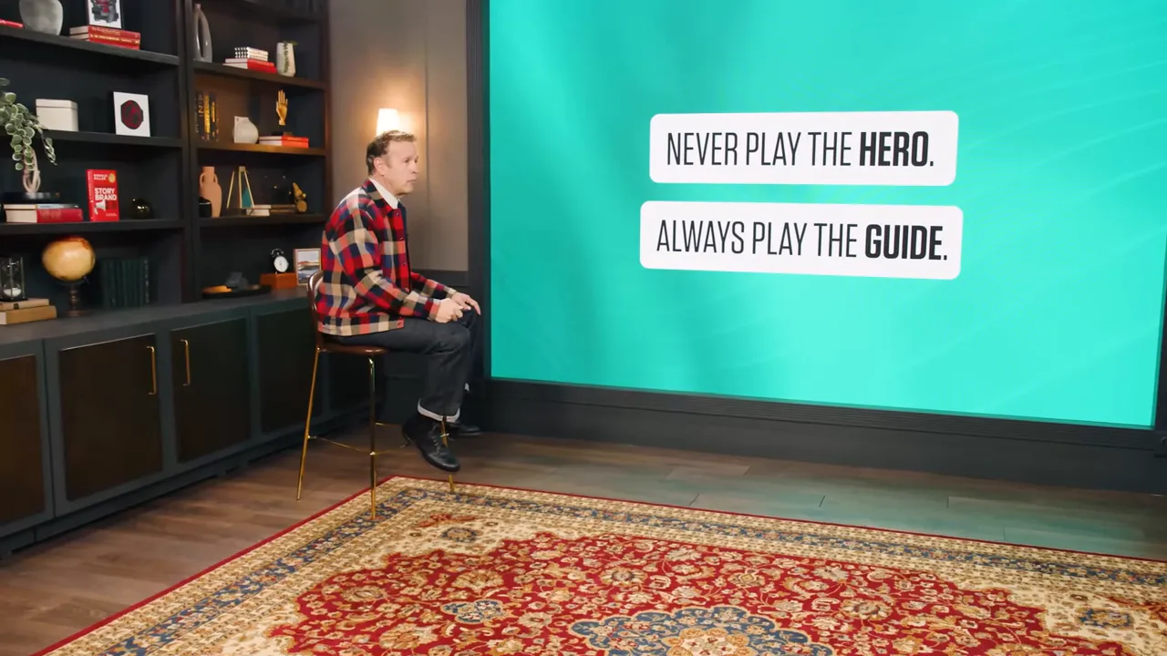 Presenter seated beside a large screen that reads 'Never play the HERO. Always play the GUIDE.' in a studio with a patterned rug.