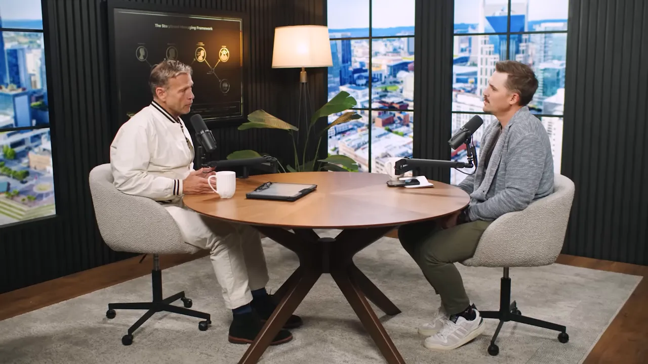 Wide studio shot of two podcast hosts seated at a round table with microphones, a tablet and a mug, city-view windows behind them.