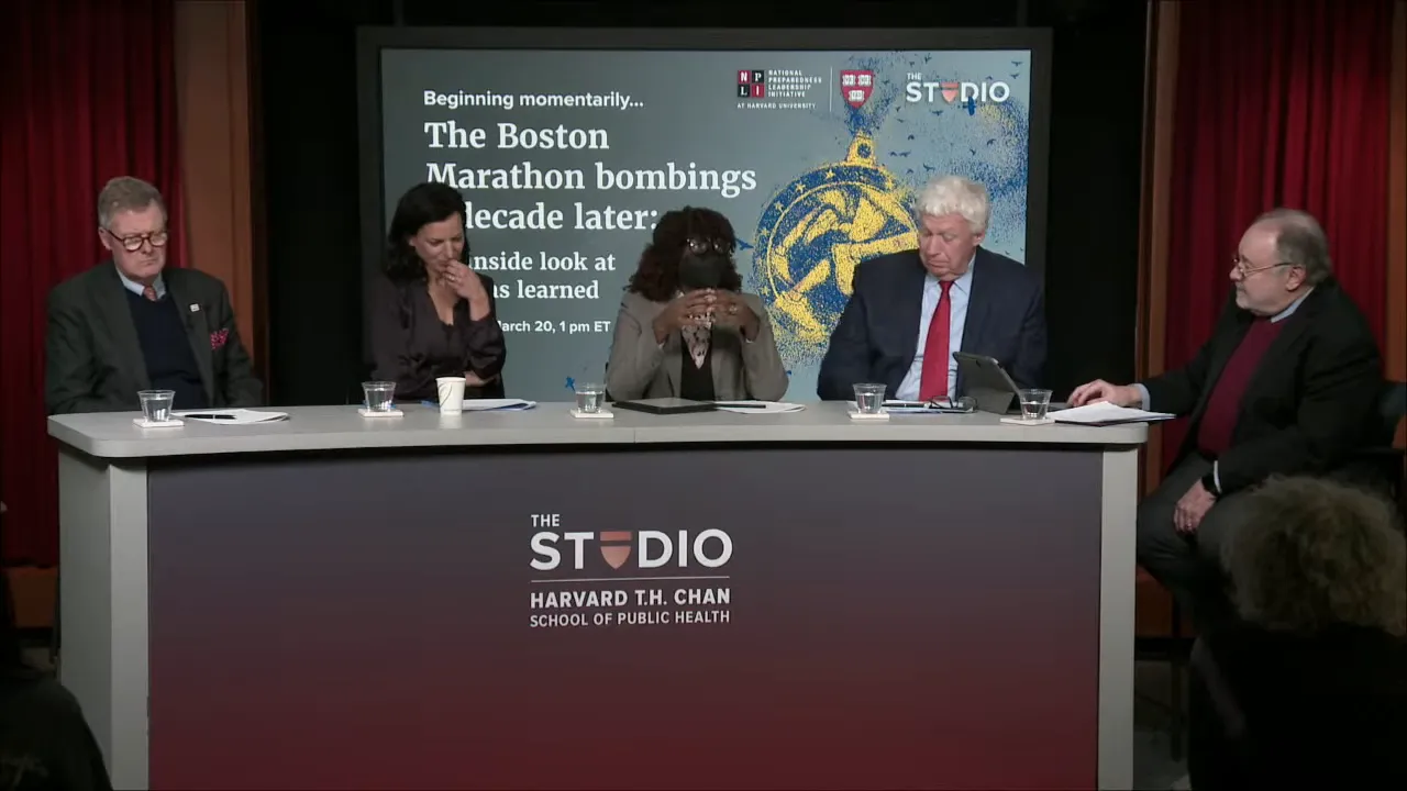 Wide shot of a multi-person panel at a conference table in front of a presentation screen reading 'The Boston Marathon bombings decade later'.