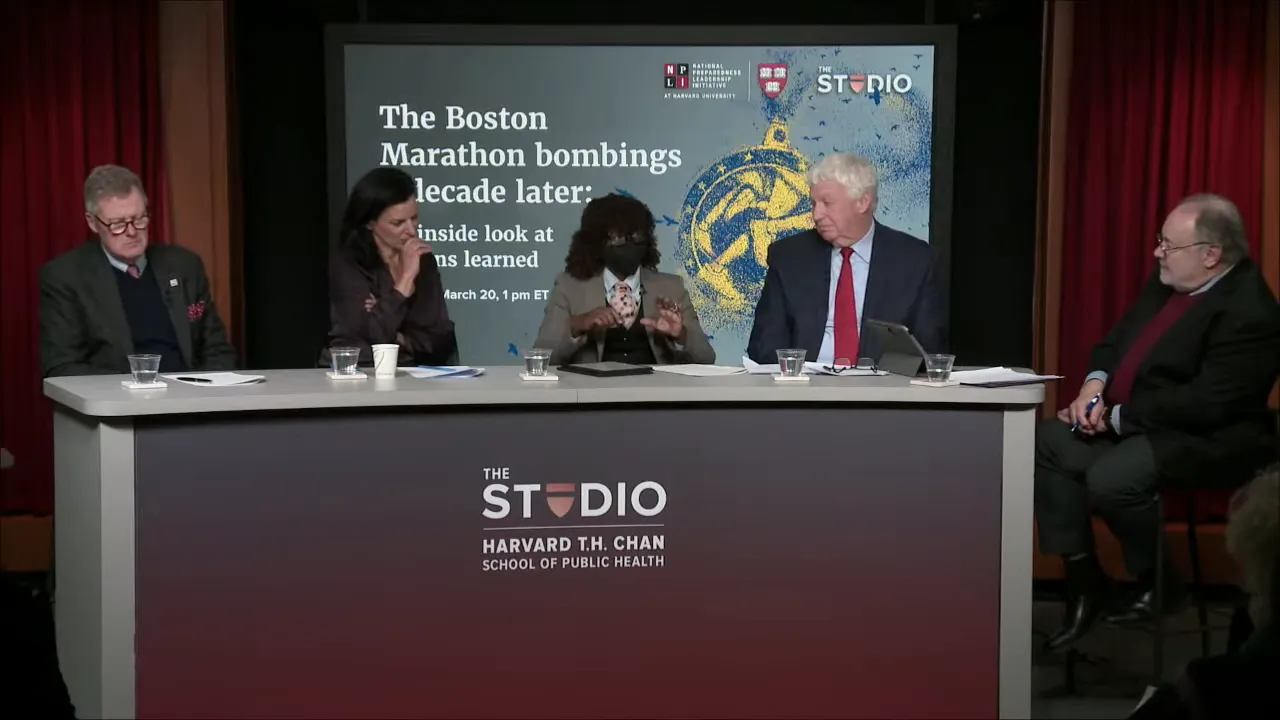 Wide shot of a multi-person panel at a table with a presentation screen reading 'The Boston Marathon bombings decade later' behind them.