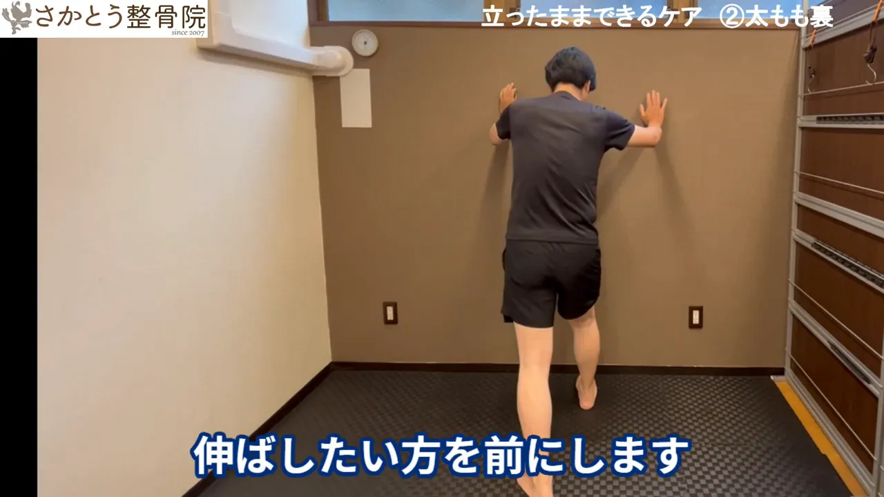 壁に両手をつき、伸ばしたい側の足を前にしてハムストリングス（太もも裏）を伸ばす準備を示す全身ショット。