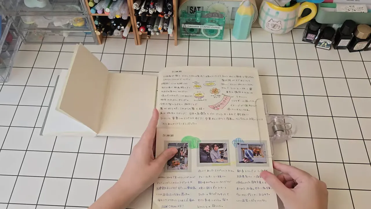 格子柄の机上で手でノートを開き、右側の透明クリップがページをしっかり押さえている写真。中の手帳ページや貼り写真が見える。