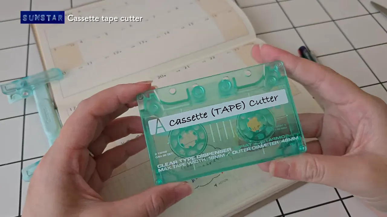 手に持った透明グリーンのカセット型マスキングテープカッターのクローズアップ（ラベルに「Cassette (TAPE) Cutter」と表示）