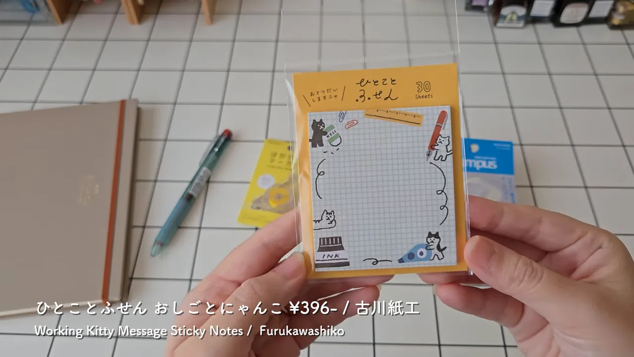 手で持たれた猫柄のメッセージ付箋パッケージ（古川紙工）のクローズアップ、背景に手帳とペン