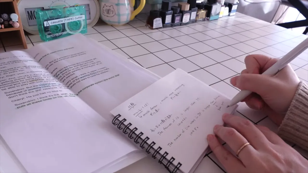 開いたミニメモに右手でペンを走らせている手元の写真。スパイラル綴じのノートがはっきり見え、デスク周りの文具も写るクリアな画像