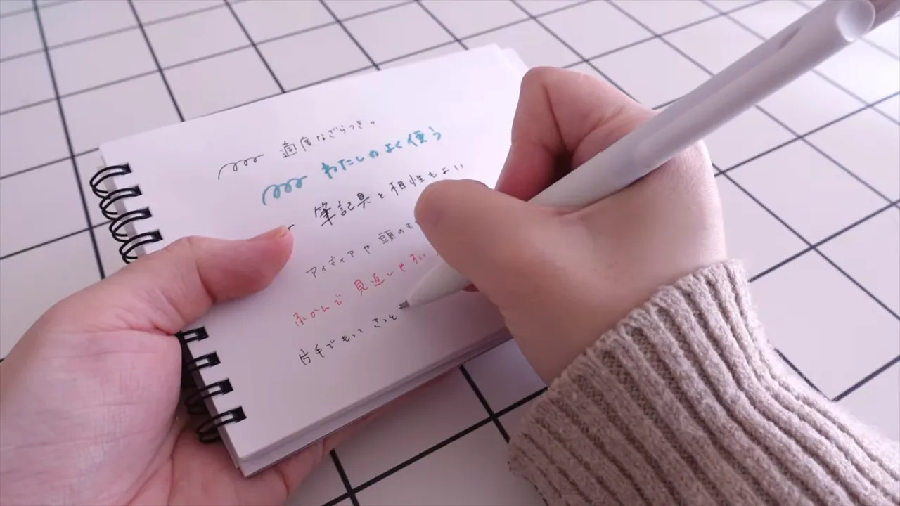 格子柄の机でミニノートに右手でペンを走らせて書き込むクローズアップ。ページと綴じ部分がはっきり見える