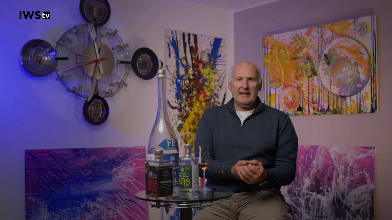 Gut fokussierte Aufnahme: Sprecher frontal, Flaschen und Verkostungsglas deutlich sichtbar