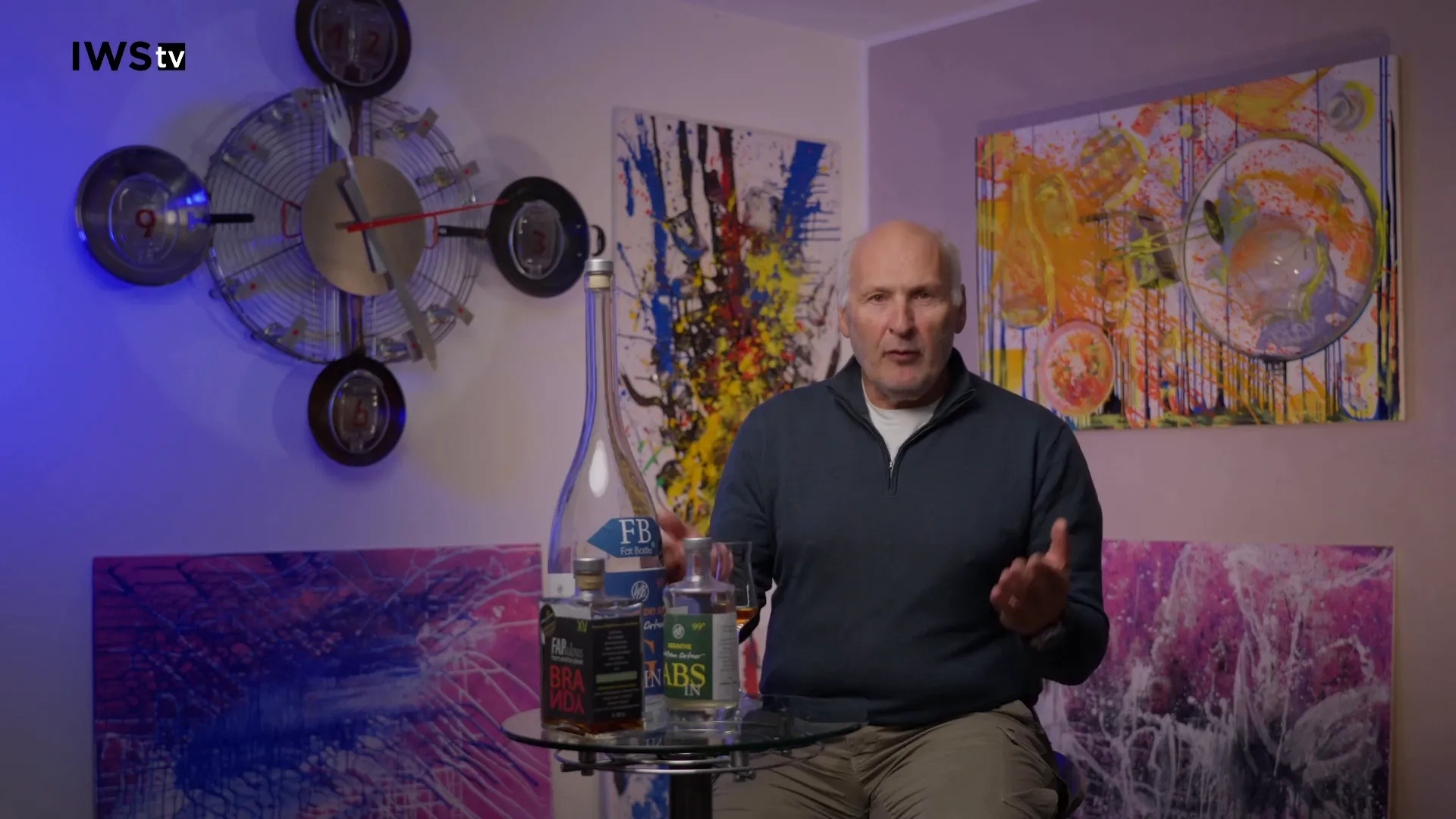 Host seated with three bottles of cachaça on a glass table, colorful paintings on the wall behind