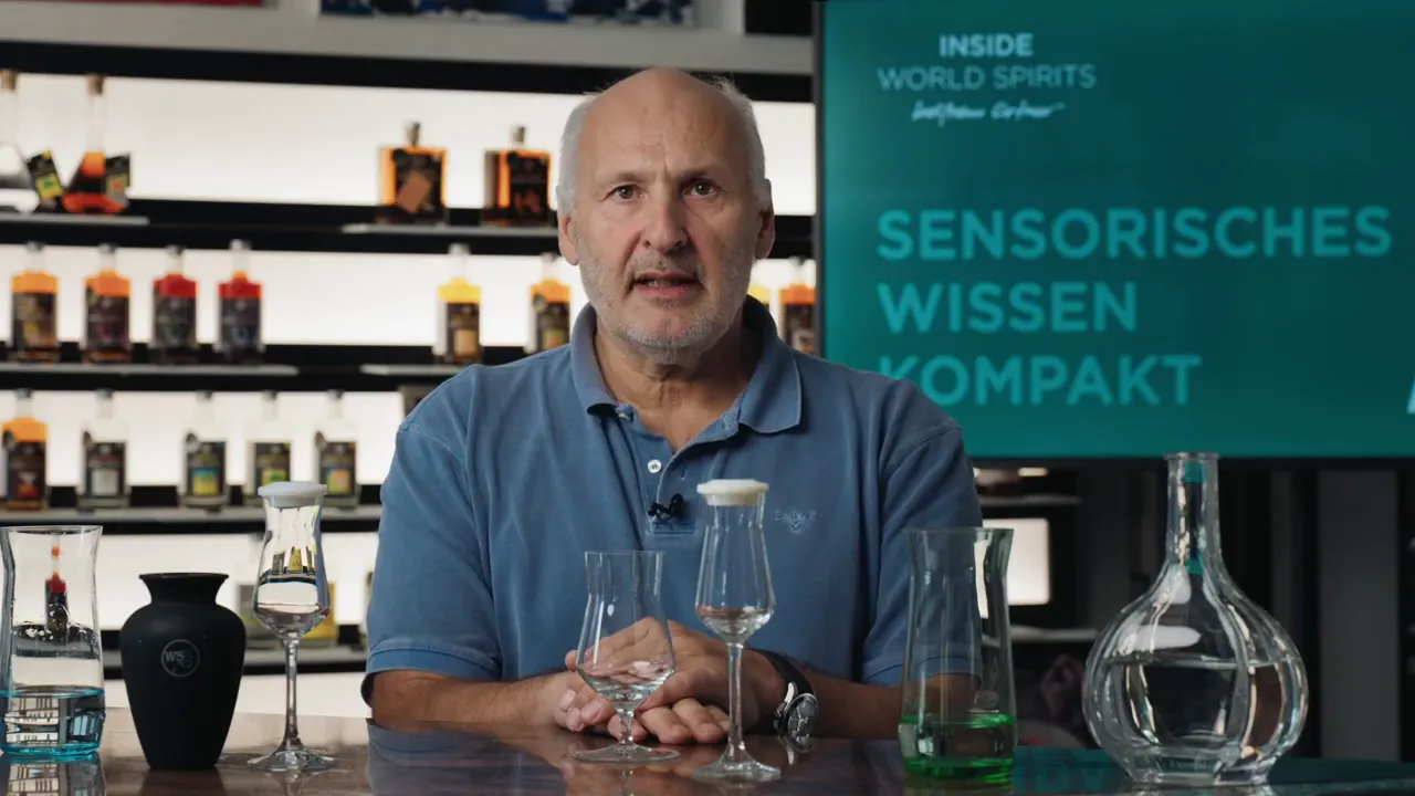Person sitzt mittig hinter einem Tisch mit mehreren Verkostungsgläsern und Karaffen; im Hintergrund ein Bildschirm mit 'Sensorisches Wissen Kompakt'.