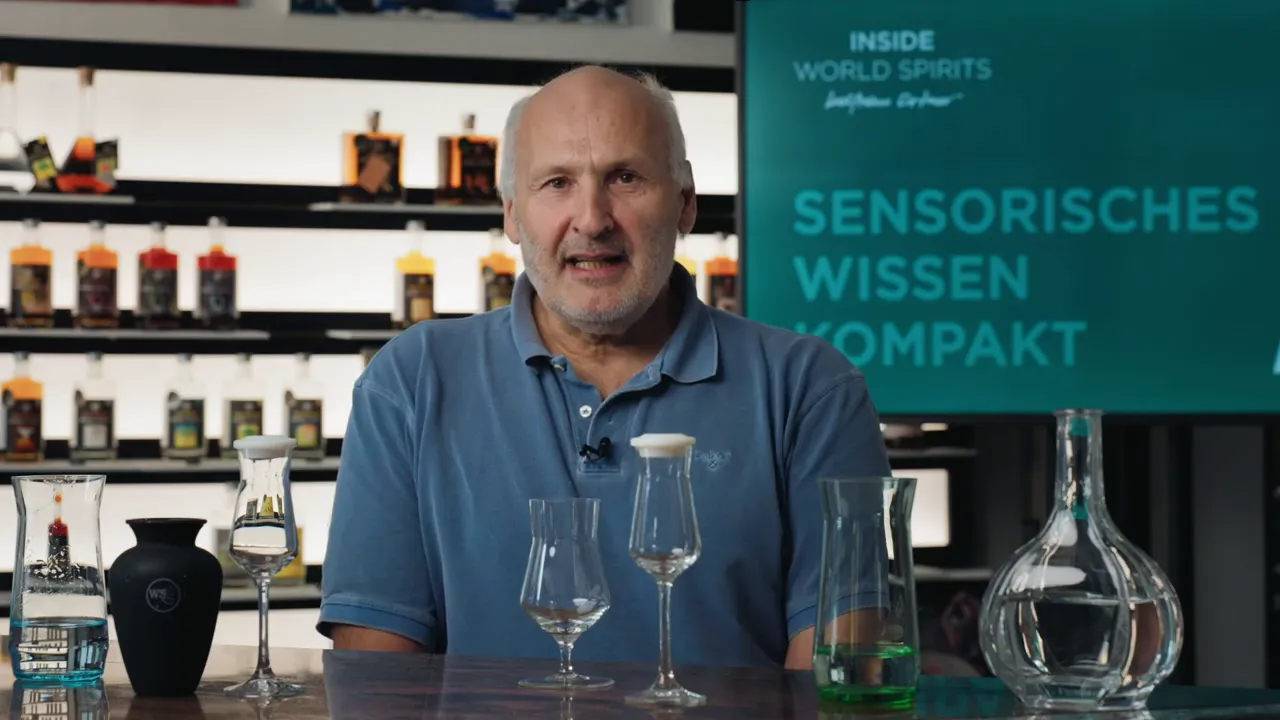 Moderator hinter einer Reihe von Verkostungs-Gläsern und einer Karaffe; im Hintergrund das Display 'Sensorisches Wissen Kompakt'.