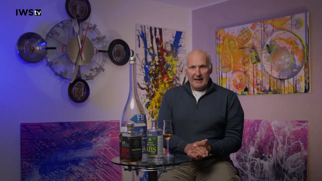 Mann sitzt neben einem kleinen Glastisch mit mehreren Flaschen und einem Verkostungsglas, bunte Bilder im Hintergrund