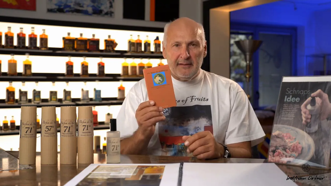 Well-lit view of Parfum de Vie bottles and cardboard tubes on a counter, the presenter holding a booklet and a poster showing a hand spraying food visible to the right.