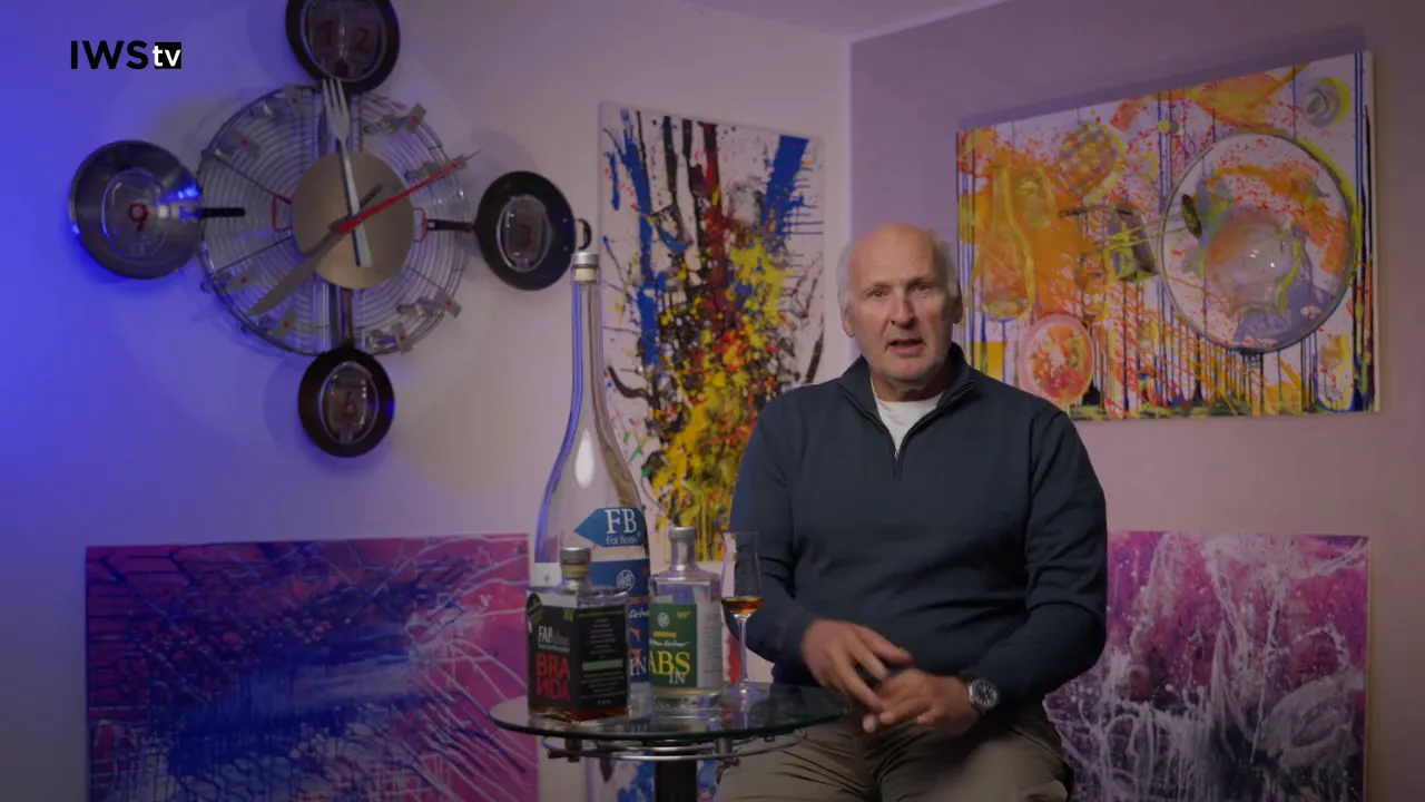 Sprecher sitzt neben mehreren Spirituosenflaschen und einem Glas auf einem kleinen Tisch, bunte Bilder im Hintergrund