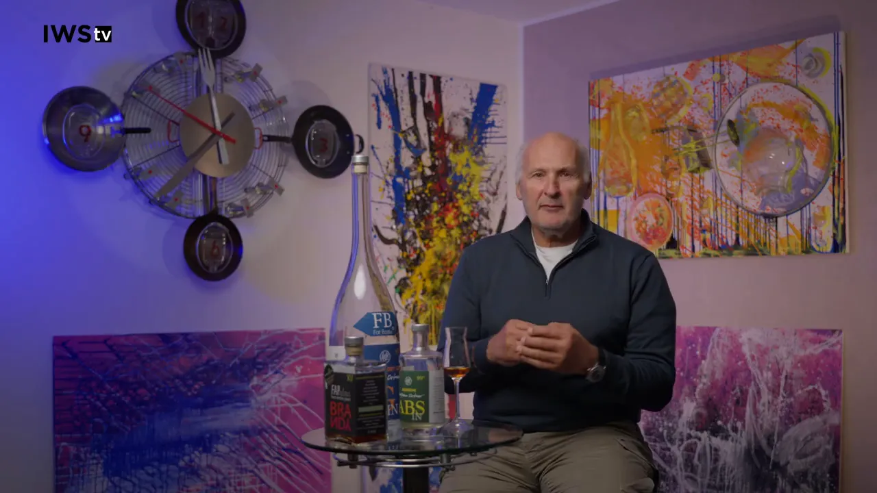 Moderator frontal, deutlich sichtbare Flaschen und Glas auf Glastisch, bunte Bilder im Hintergrund