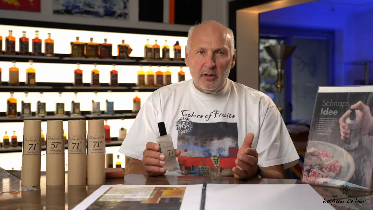 Moderator hält Flasche Parfum de Vie 71 auf dem Tisch; Verpackungen und Regal mit Destillaten im Hintergrund