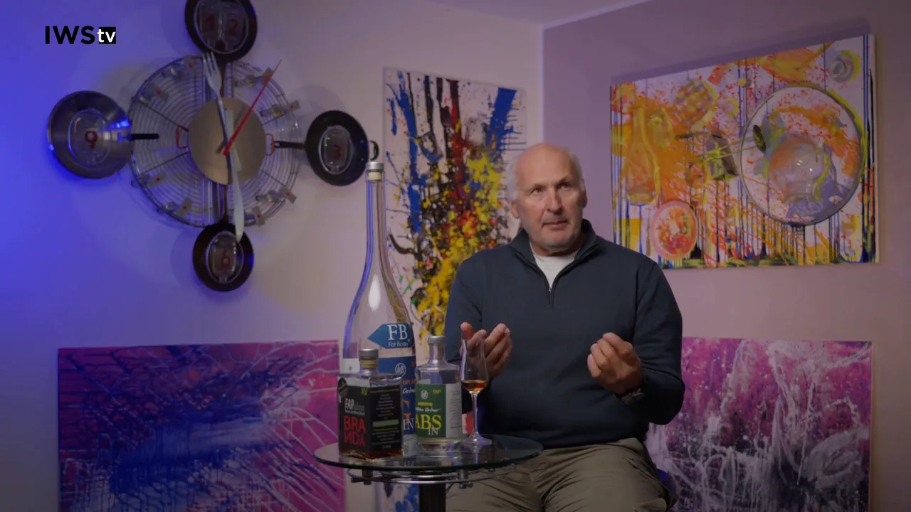 Bottles and a tasting glass with amber spirit on a small table while the speaker gestures during an interview