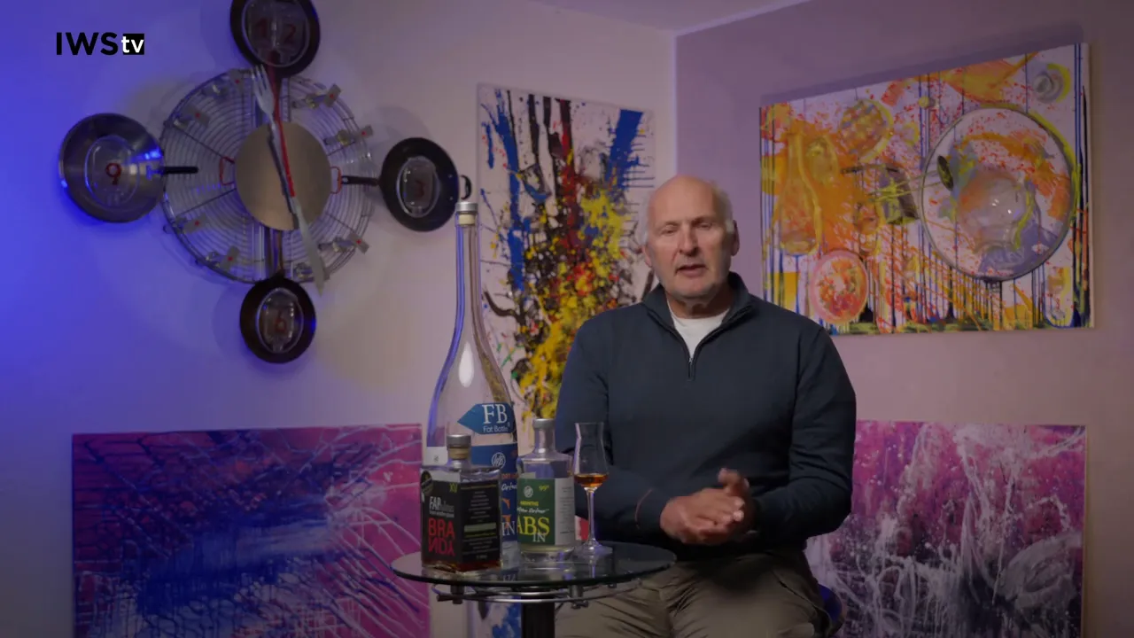 Wide shot of presenter seated beside a small table with multiple calvados bottles and a tasting glass, colorful artwork in the background.