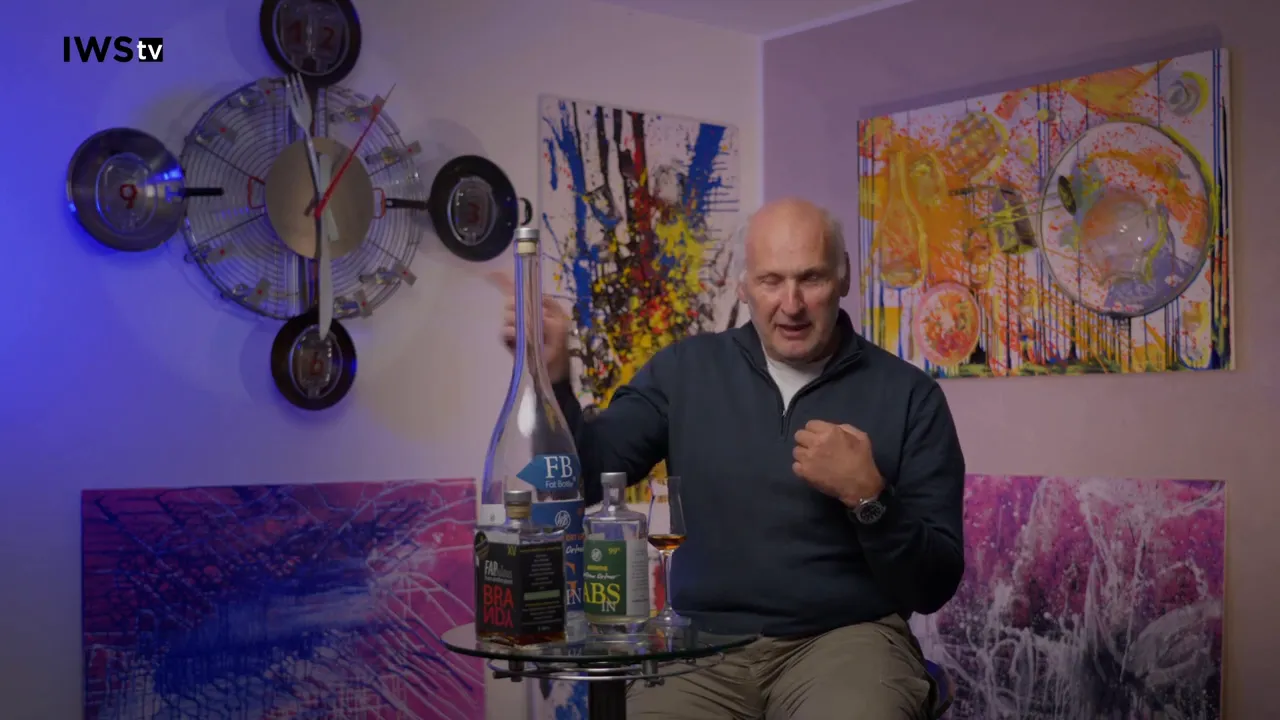 Host seated with bottles of calvados and a tasting glass on a small round table, colorful artwork in the background