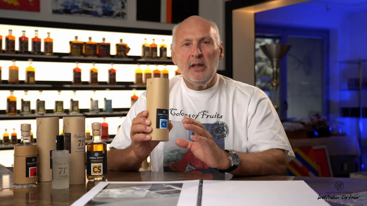 Moderator präsentiert eine zylindrische 71%-Essenzverpackung in der Hand, mehrere Essenzflaschen links auf dem Tisch, Regal mit Flaschen im Hintergrund