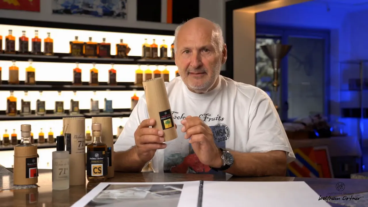 Wide shot of presenter at a table with several spirit bottles and a cylindrical essence package, well lit and in clear focus.