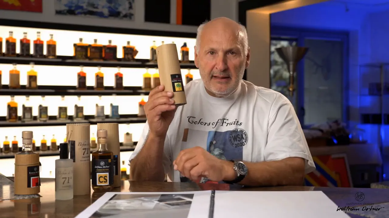 Presenter seated at a tasting table holding a cylindrical essence tube with several small essence bottles and backlit shelves visible behind