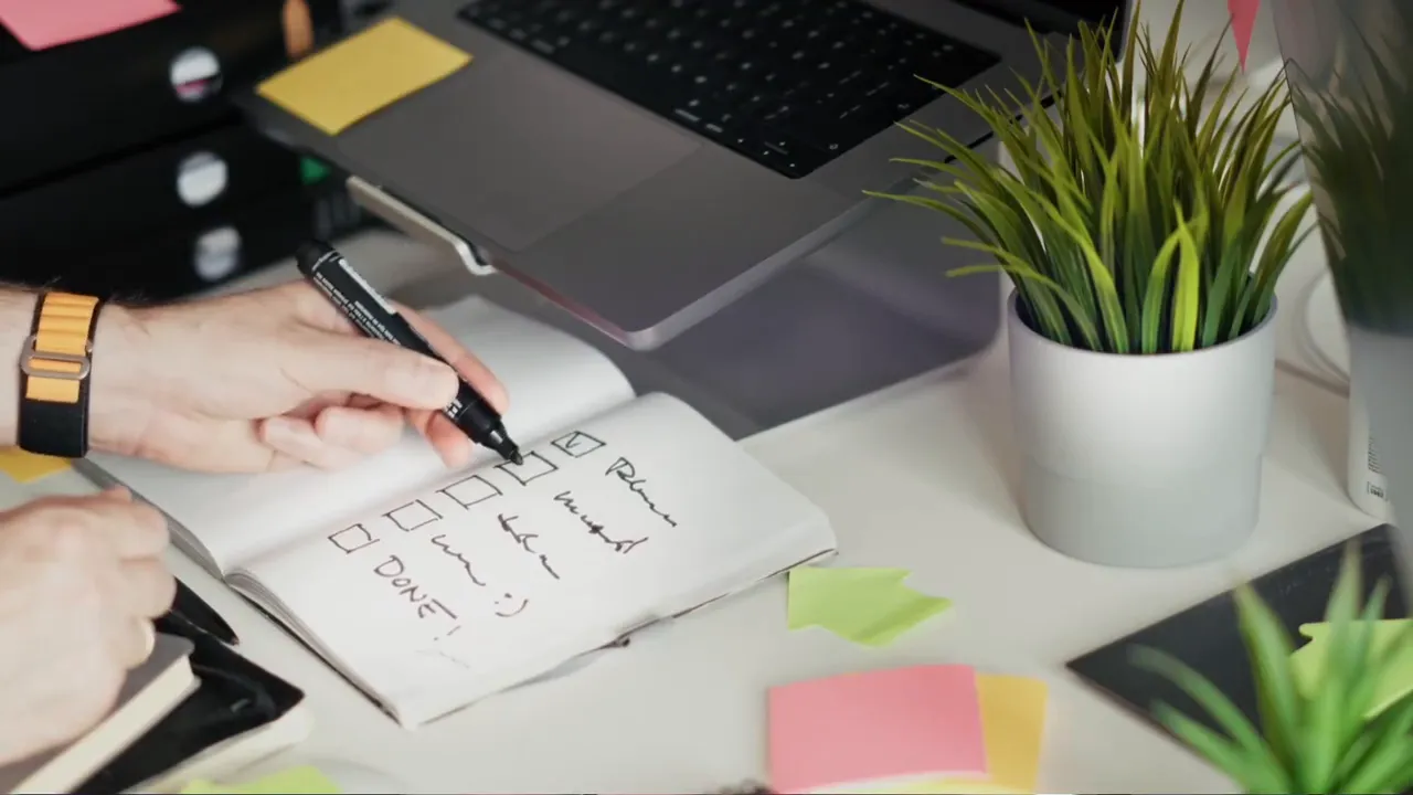 Nahaufnahme eines offenen Notizbuchs mit handschriftlichen Aufgaben und einer markierten Checkbox neben Laptop und einer Topfpflanze.