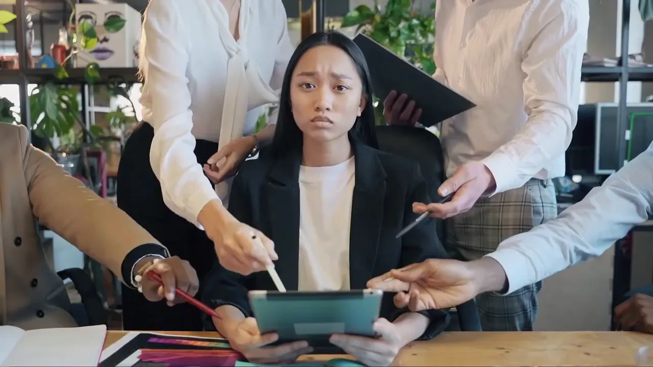 Person sitzt am Schreibtisch, mehrere Hände zeigen auf Tablet – symbolisiert Überforderung durch zu viele Inputs