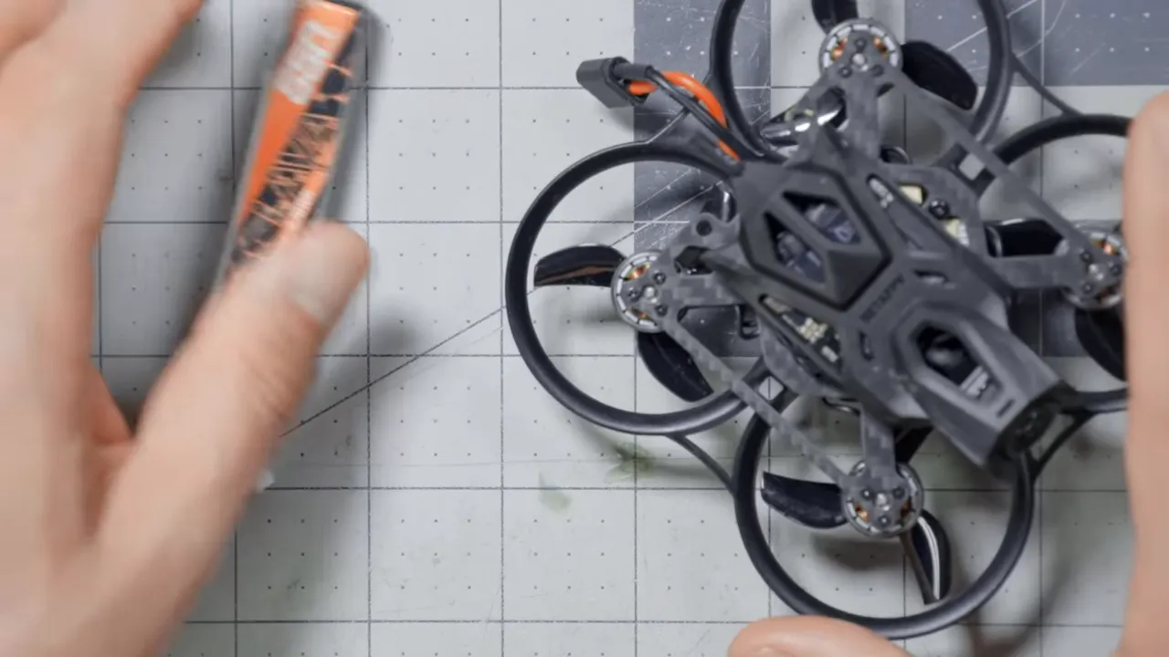 Top-down view of a BetaFPV Pavo Pico II on a work mat with hands and a battery, showing prop guards and frame detail