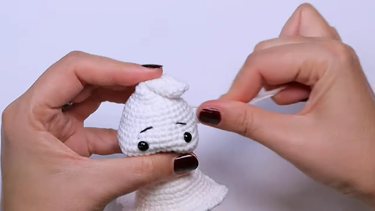 Hands holding a completed white crochet ghost with black eyes, embroidered eyebrows and smile, a curled tip, and a flared skirt