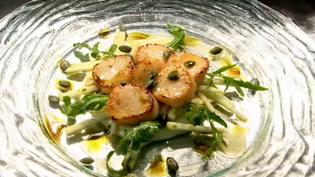 Top-down view of five golden seared scallops arranged on julienned apple and rocket with pumpkin seeds and a light drizzle on a rippled glass plate.