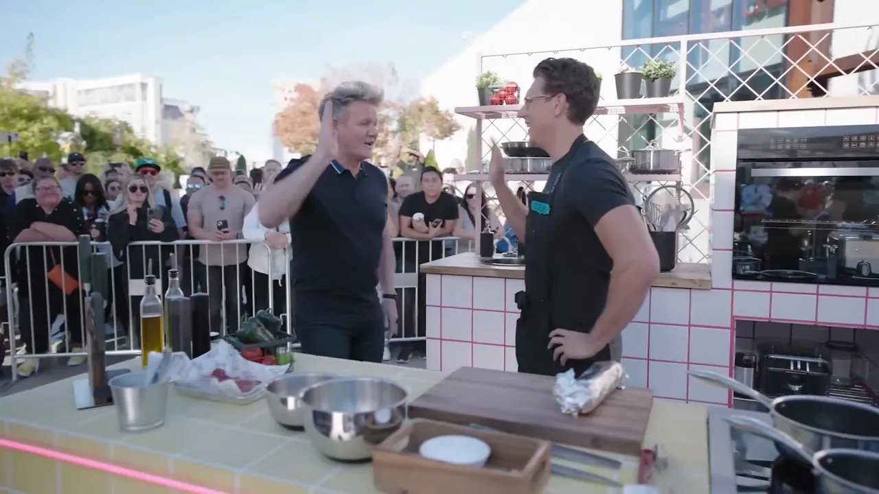 Two chefs share a celebratory high-five on an outdoor cooking stage with prep bowls, a cutting board and the audience visible behind them.