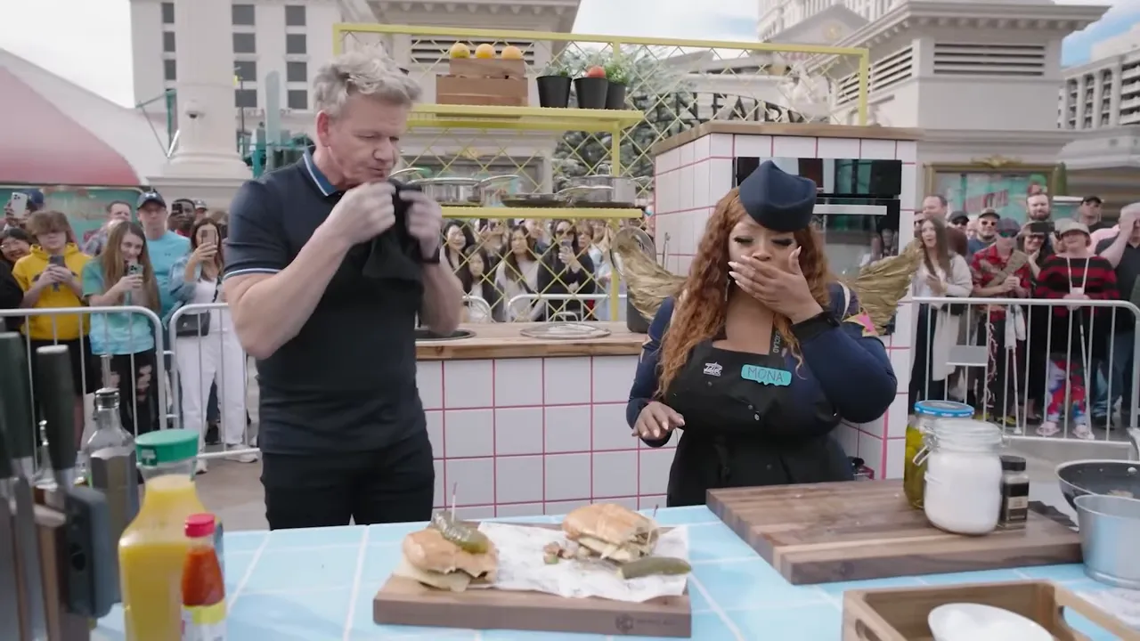 Two finished sandwiches on cutting boards at the front of a cooking station with two cooks and an outdoor crowd behind them.
