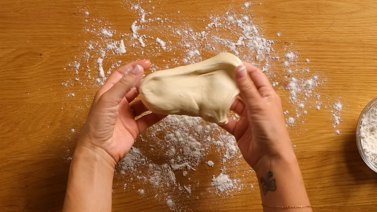 Two hands stretching a piece of pizza dough over a floured wooden surface, showing a silky, elastic texture.