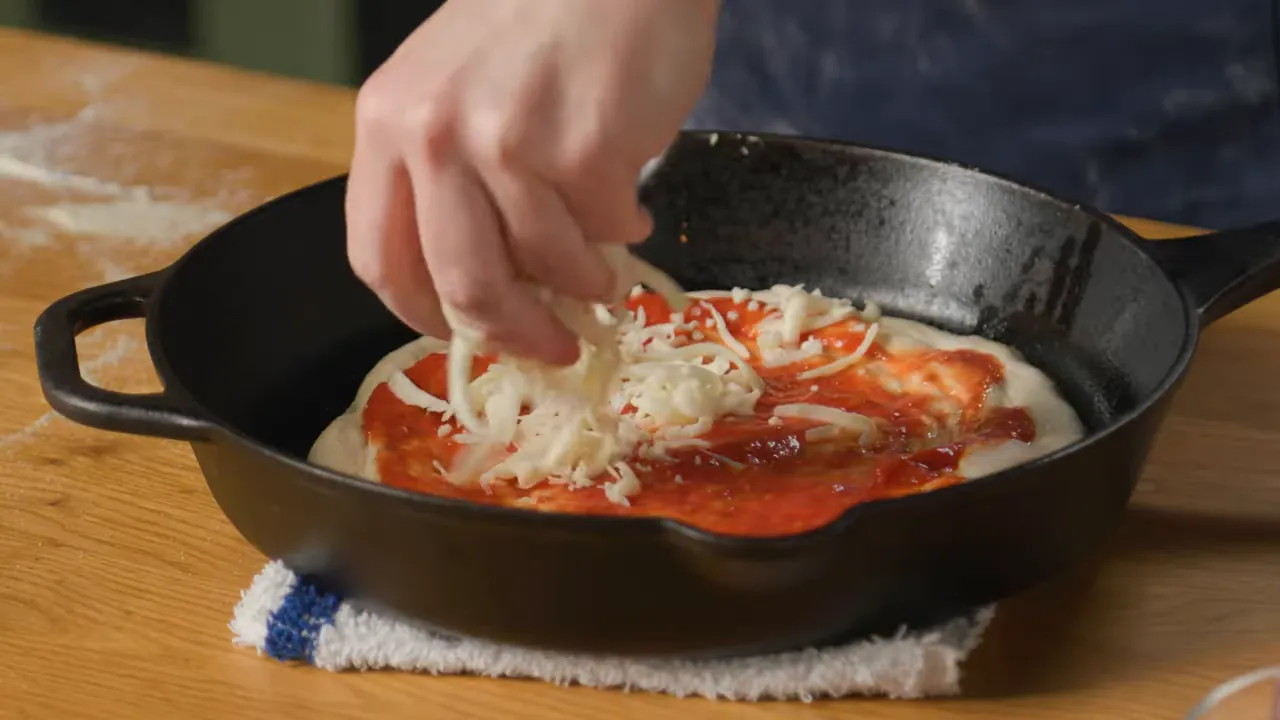 hand sprinkling shredded mozzarella over sauced pizza in a cast iron skillet