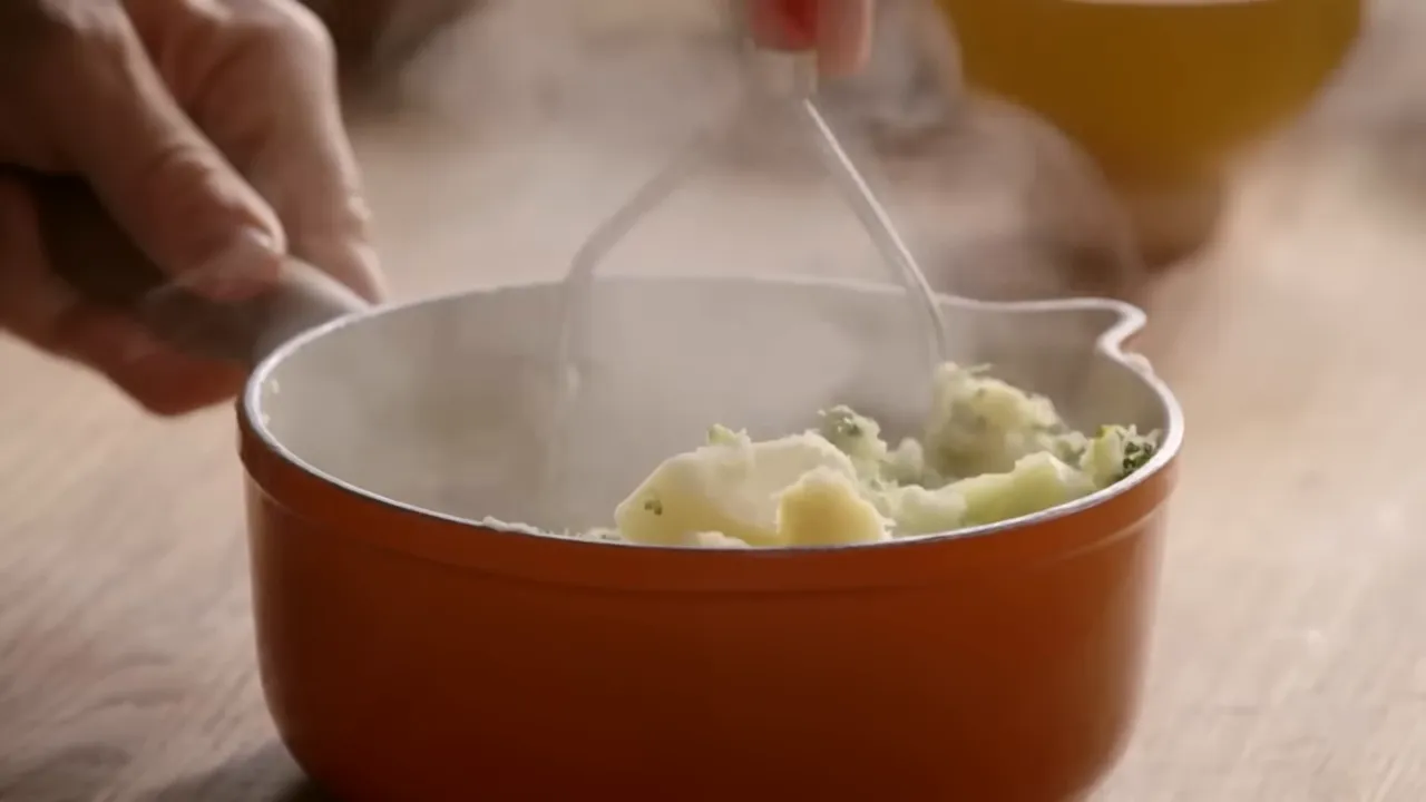 close-up of a potato masher working steaming broccoli and potatoes in an orange pot