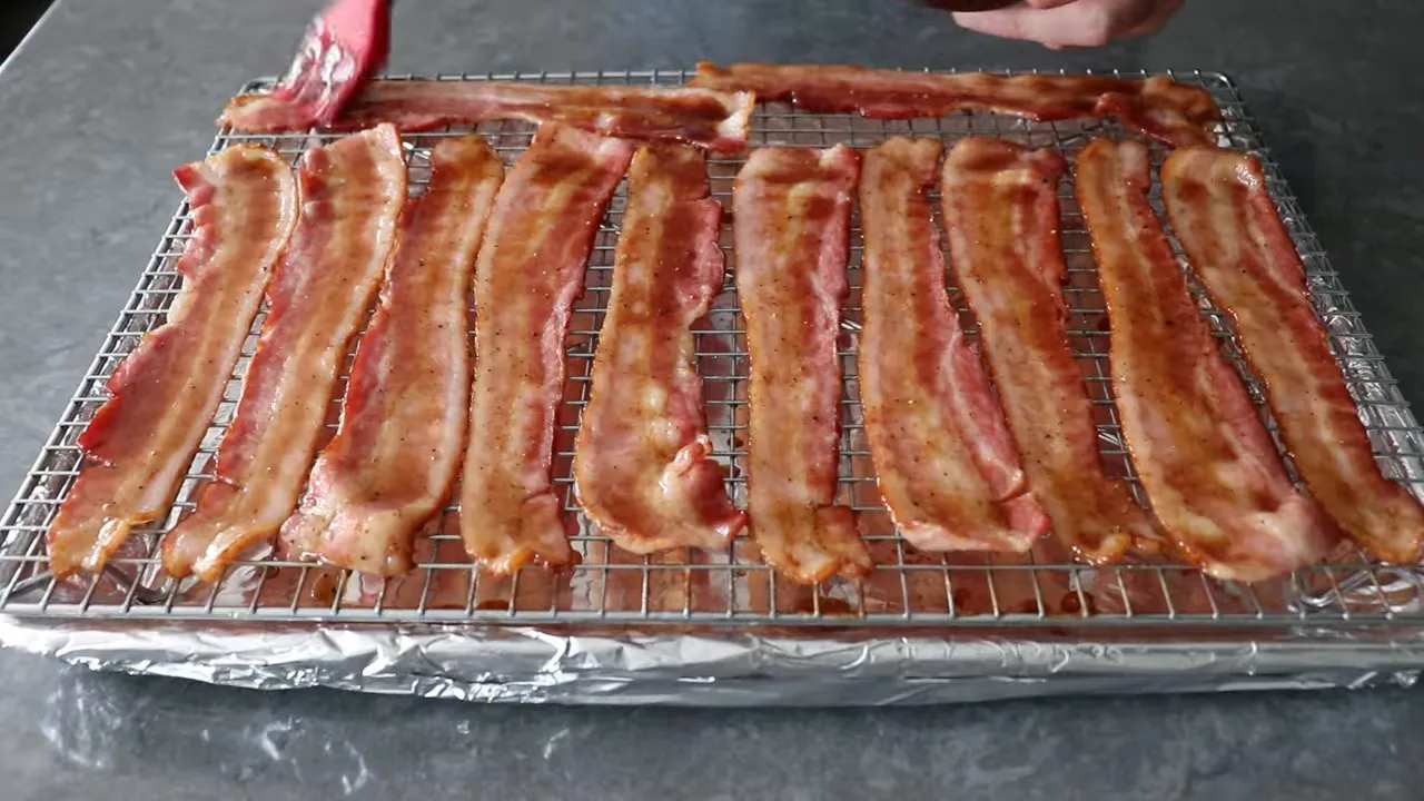 Brushing brown sugar and barbecue glaze onto par‑baked thick‑cut bacon on a wire rack