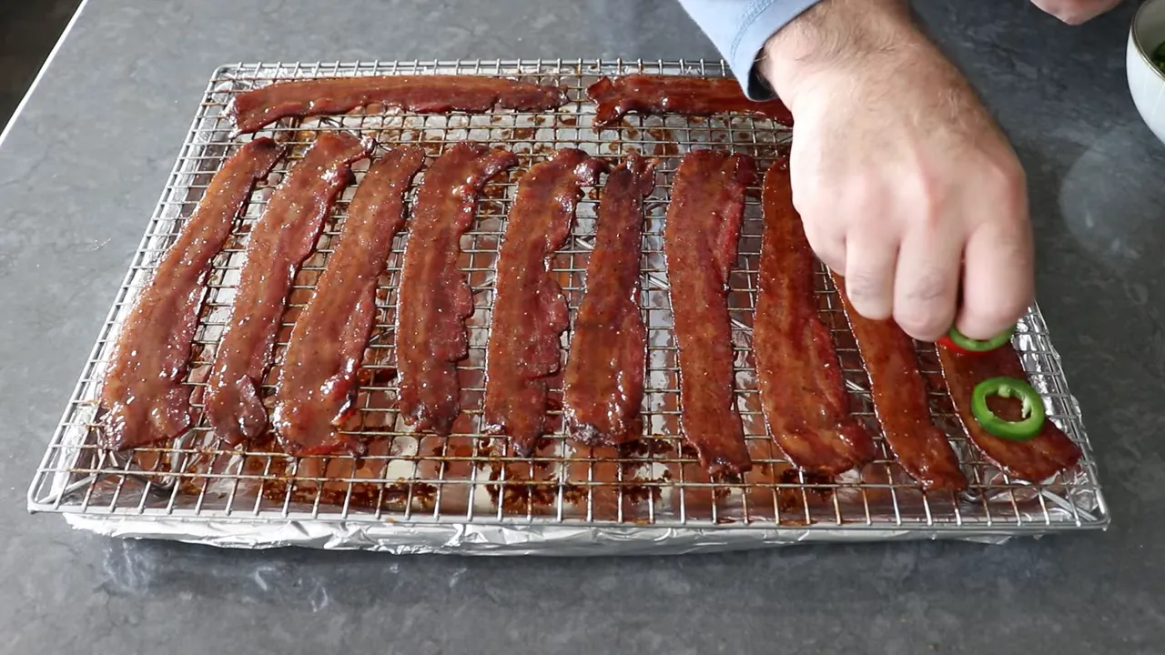 Hand placing a jalapeño ring on a glazed bacon strip on a wire rack.
