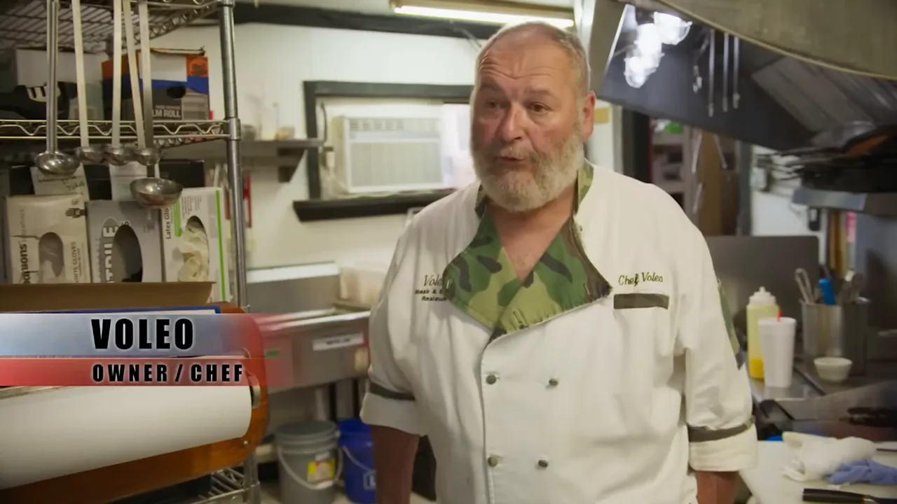 Owner-chef standing in a busy kitchen speaking, with shelving, utensils and prep equipment behind him.