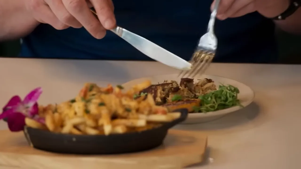 Close-up of a plated entrée with greens and a cast-iron skillet of fries in the foreground while a fork and knife finish the plate