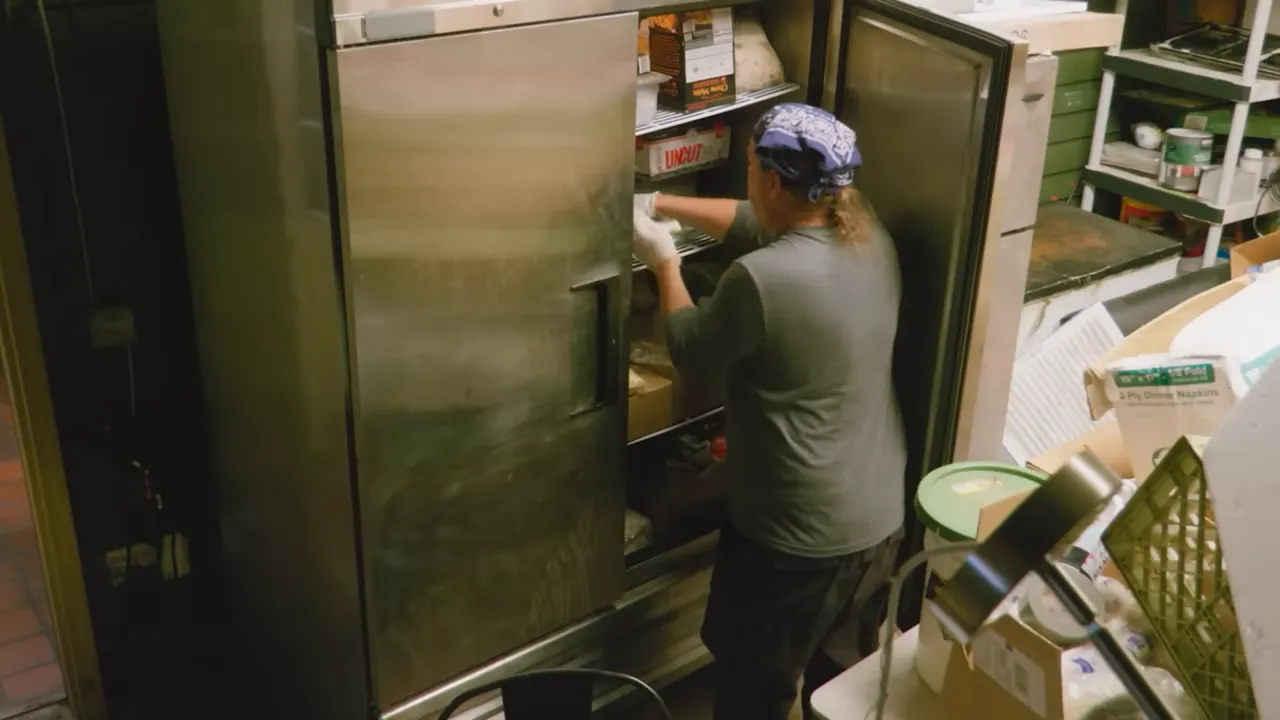Worker organizing items inside a commercial stainless refrigerator in a cluttered prep area