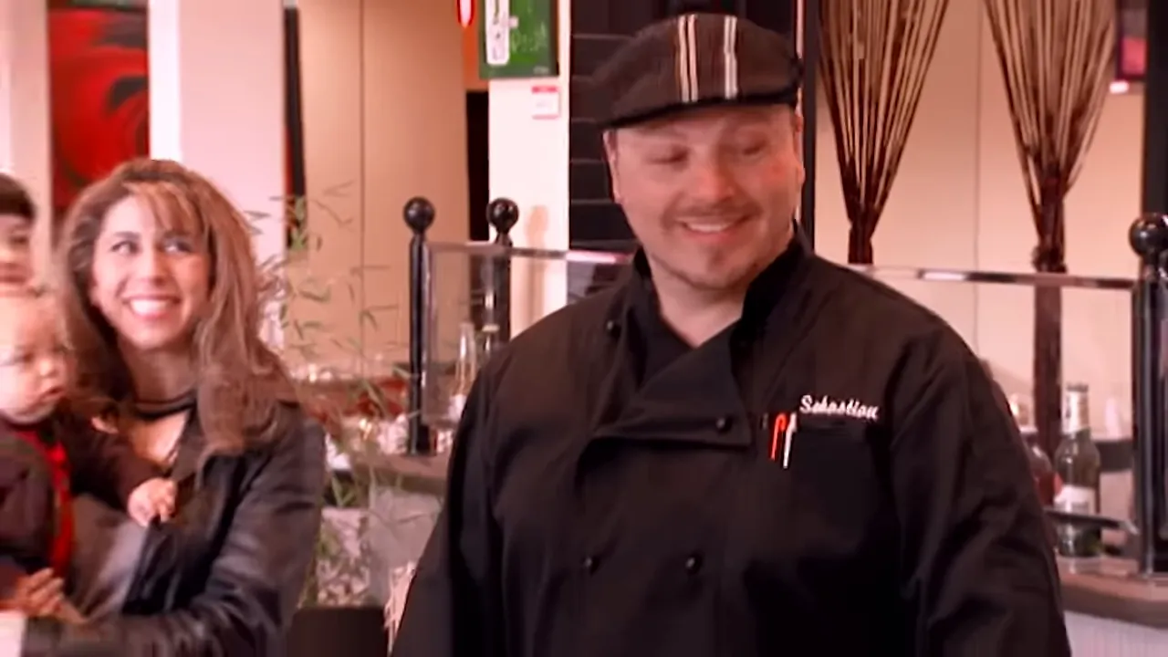 Chef in a black coat smiling with two guests/staff in a bright restaurant dining area.